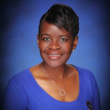 Portrait of an African American woman with short black hair, wearing a blue top and a necklace, smiling against a blue gradient background.