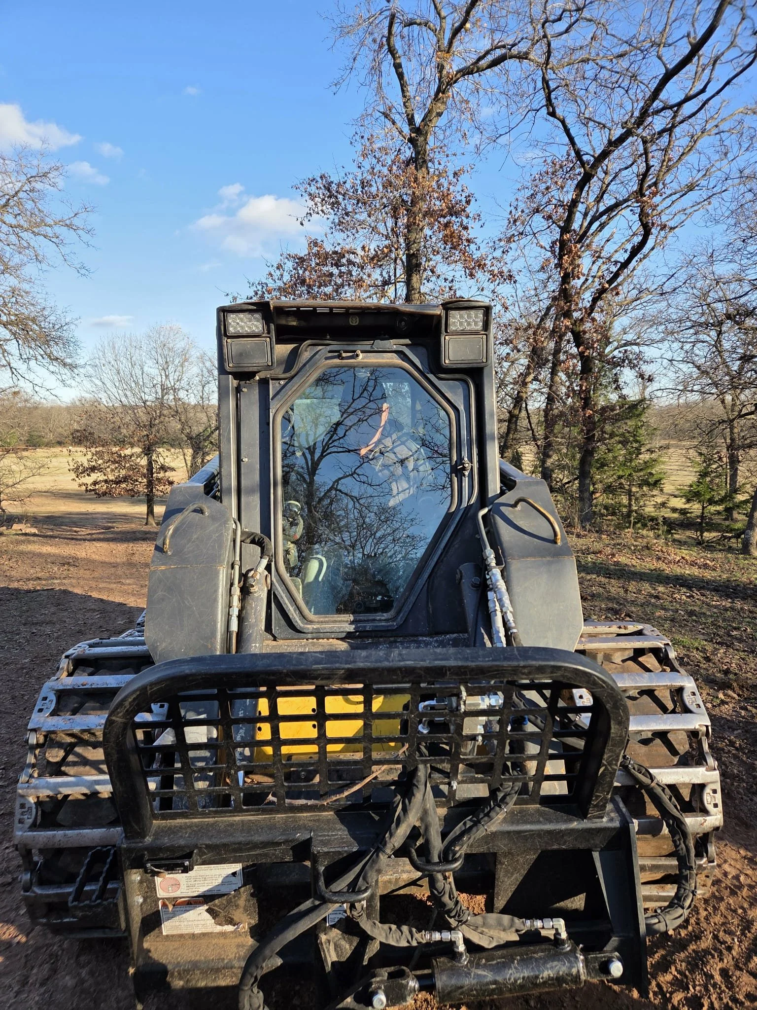A front view of a black all-terrain vehicle (ATV) or utility task vehicle (UTV) on a dirt trail in a wooded area with mostly bare trees and a clear blue sky.