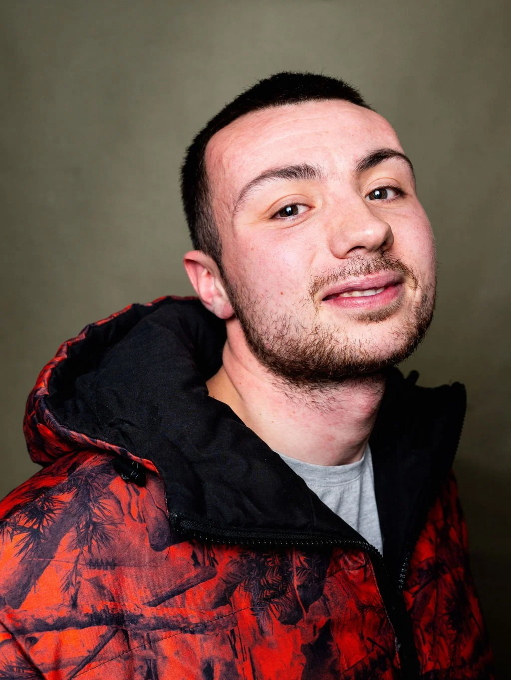 A young man with short dark hair and facial hair, wearing a black and red jacket and a gray shirt, smiling slightly against a plain background.