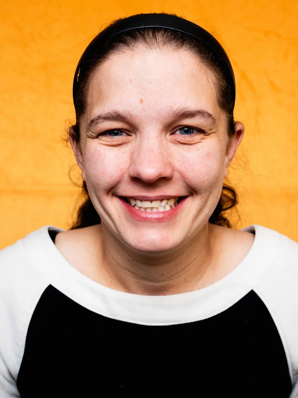 Close-up portrait of a smiling woman with blue eyes wearing a black headband and black-and-white shirt, set against a yellow background.