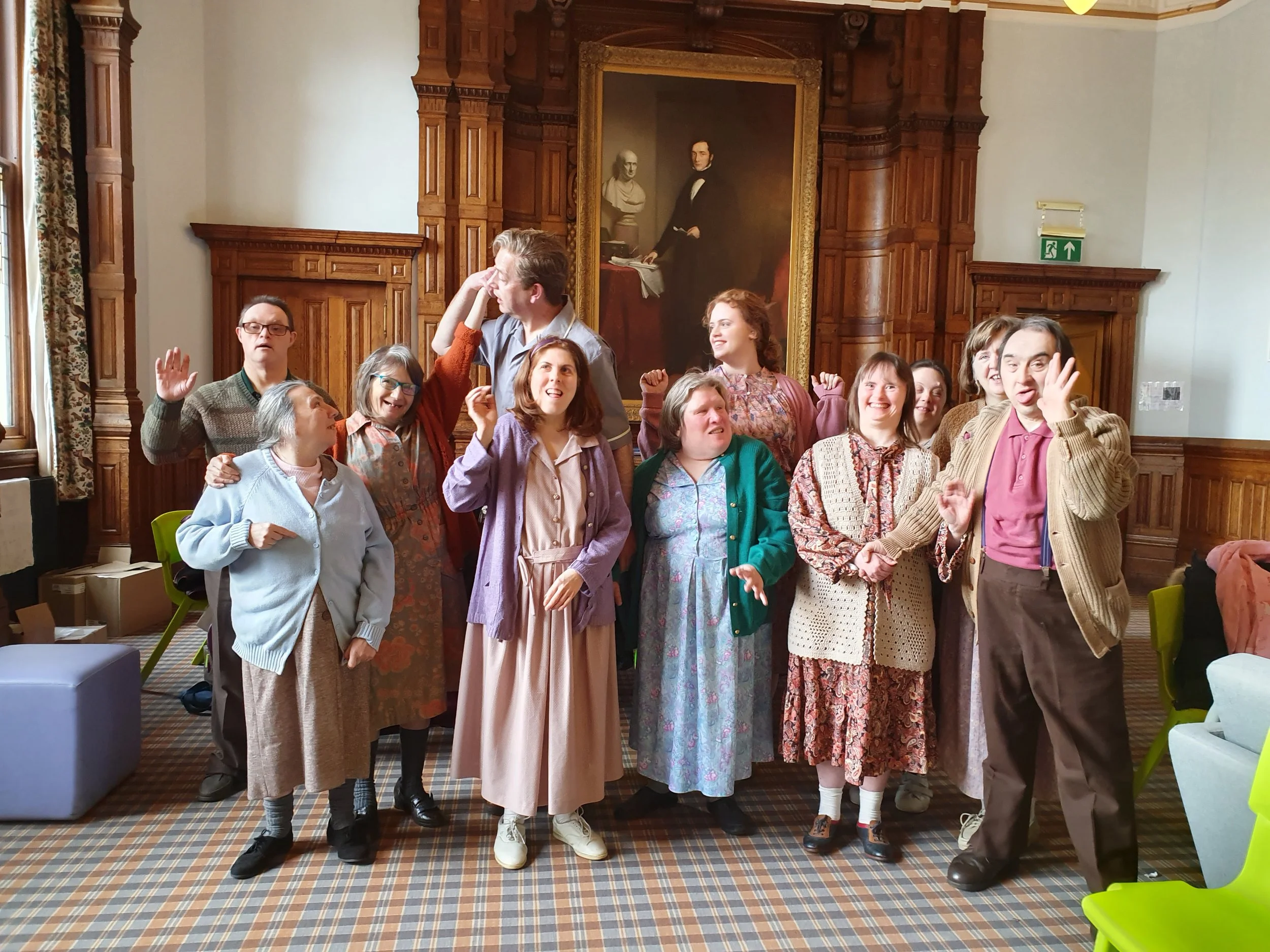 A group of eleven people, mostly women, dressed in vintage clothing, posing and making funny gestures in a room with wooden paneling and a large portrait painting in the background.