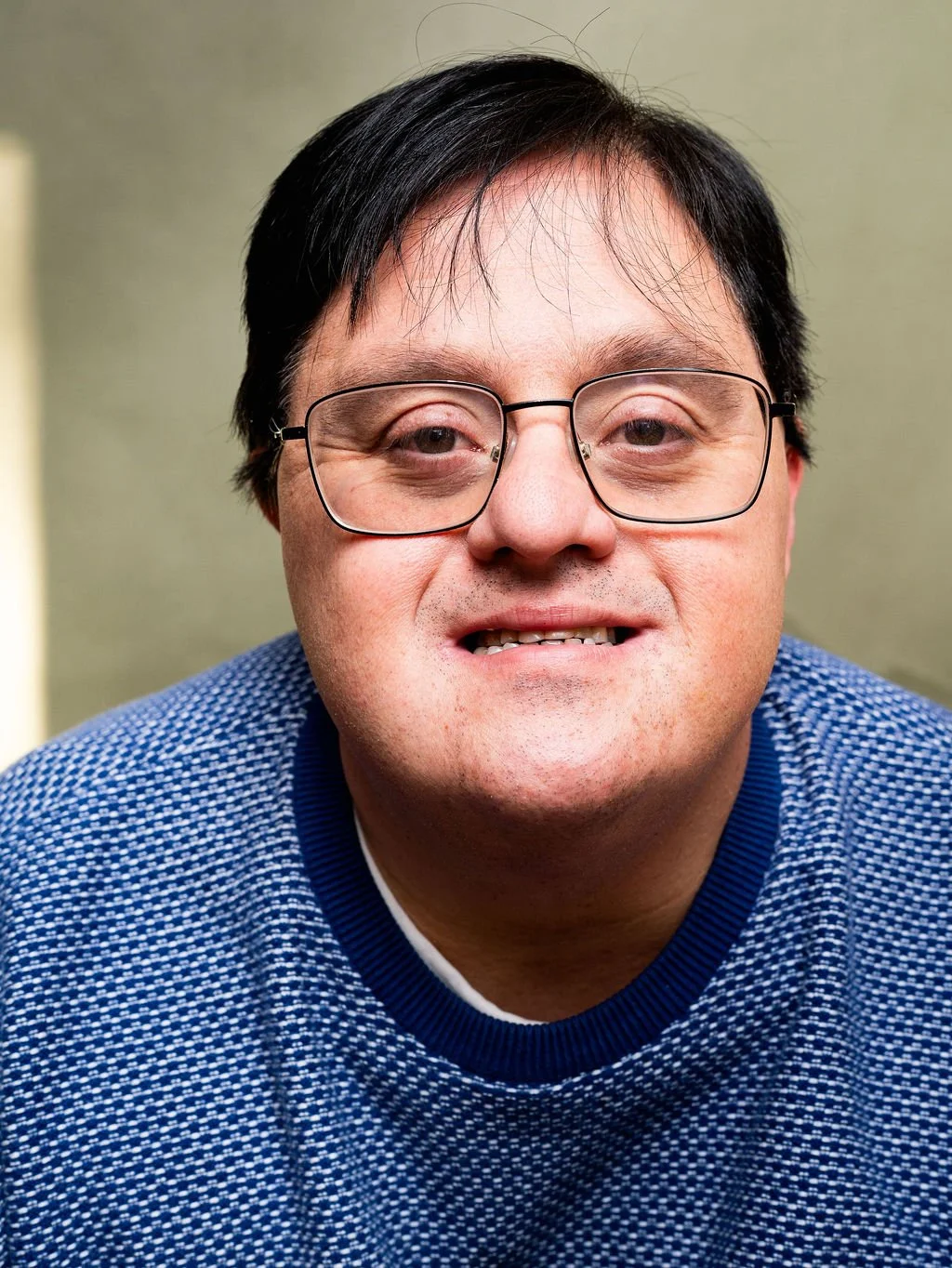 Close-up portrait of a man with black hair, glasses, and wearing a blue patterned sweater, smiling slightly, in front of a neutral background.