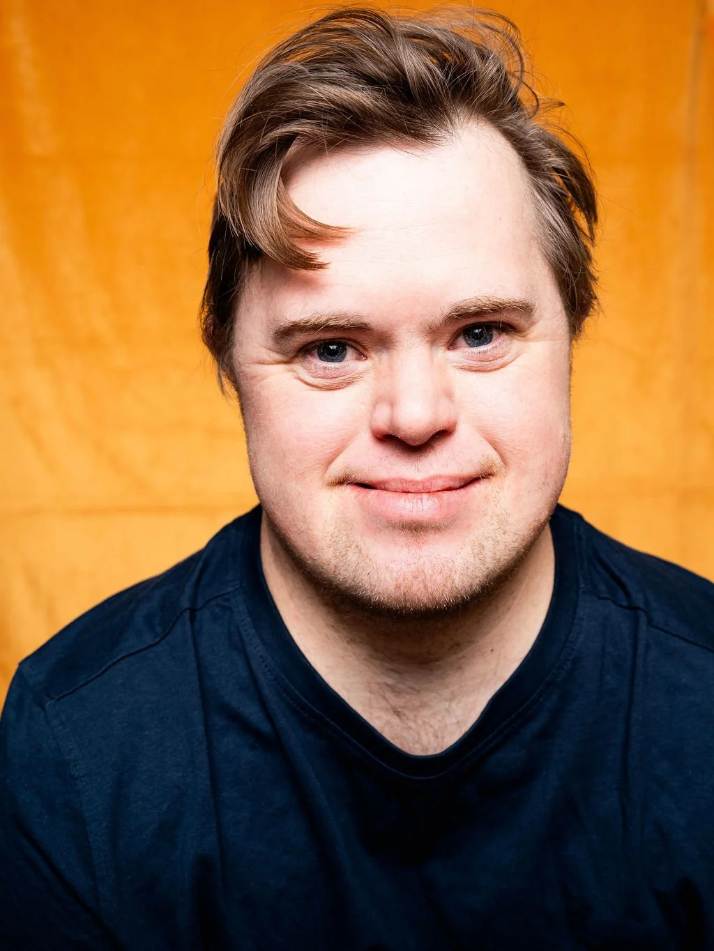 A young man with light brown hair, and blue eyes, smiling, wearing a dark blue t-shirt, standing in front of a wooden background.