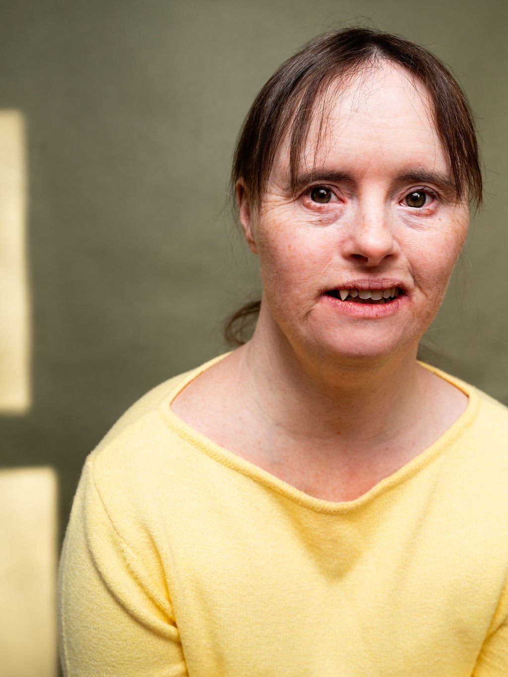 Close-up of a woman with short brown hair, wearing a yellow top, standing indoors against a green wall.