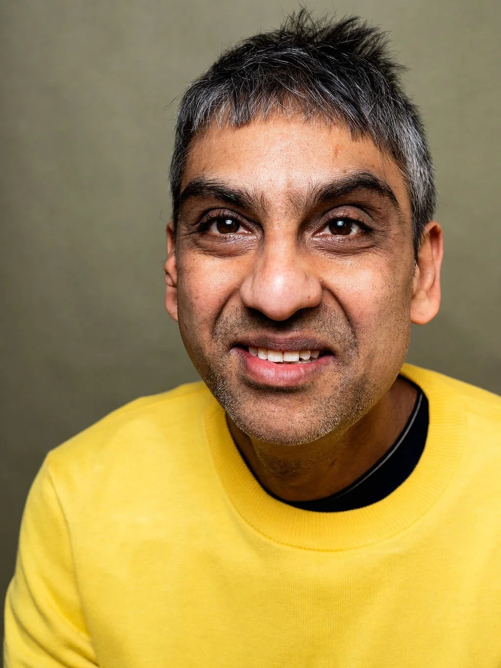 Close-up of an Asian man with grey and black hair, wearing a yellow shirt, and smiling.