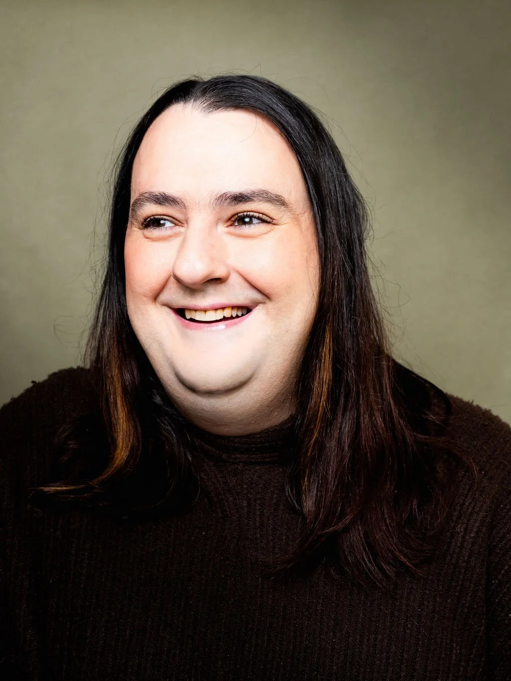 A trans woman with long dark hair, wearing a dark sweater, against a plain neutral background.