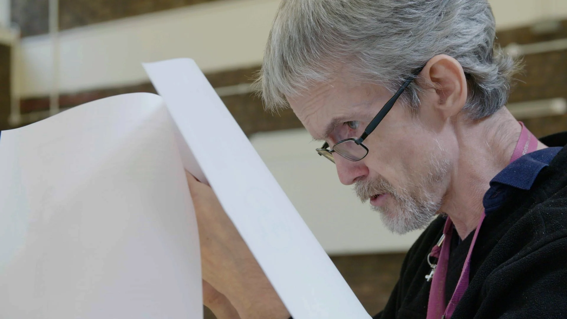 A middle-aged White man with grey hair, glasses, and a beard leaning over and intently reading a script.