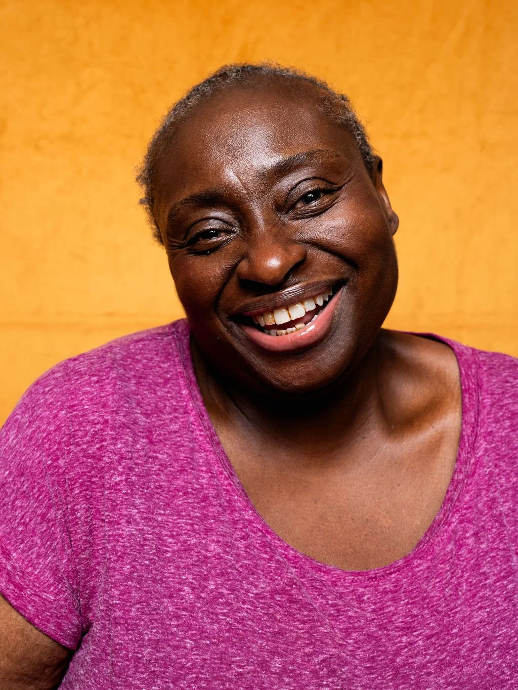 A smiling Black woman with short hair, wearing a pink shirt against a yellow background.