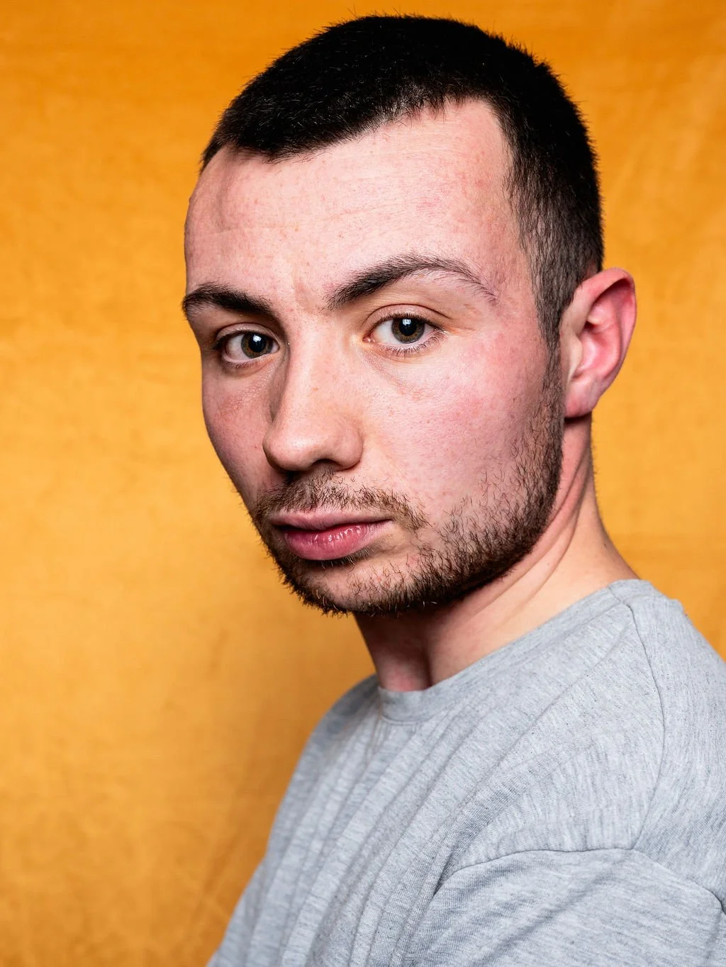 Close-up portrait of a young man with short dark hair and blue eyes, wearing a light grey shirt, against a yellow background.