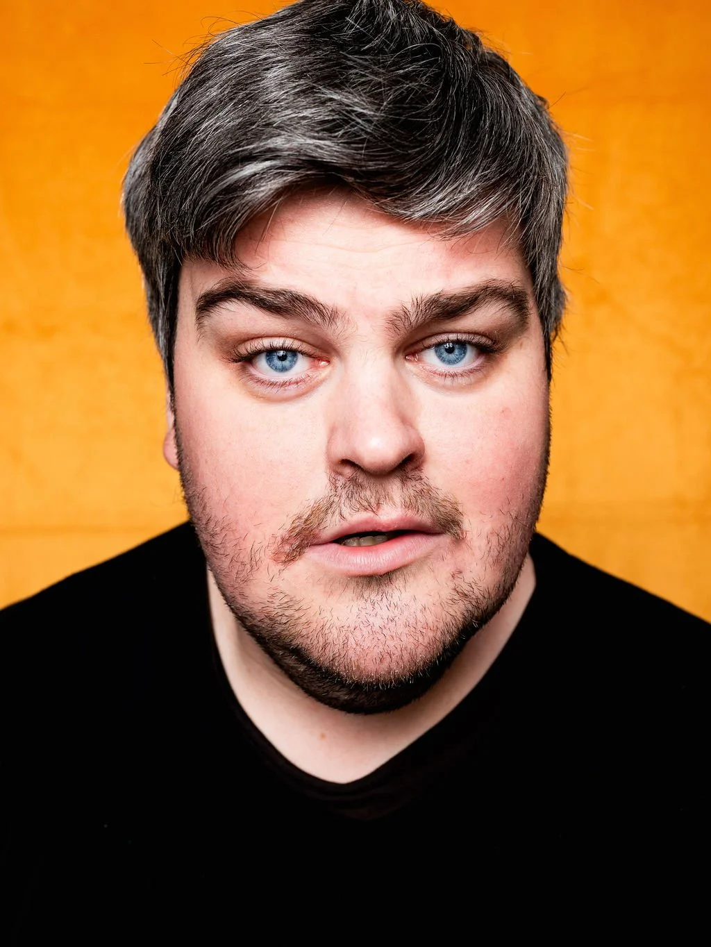 Close-up of a young White man with blue eyes, dark brown hair, and beard, wearing a black shirt, against an orange background.