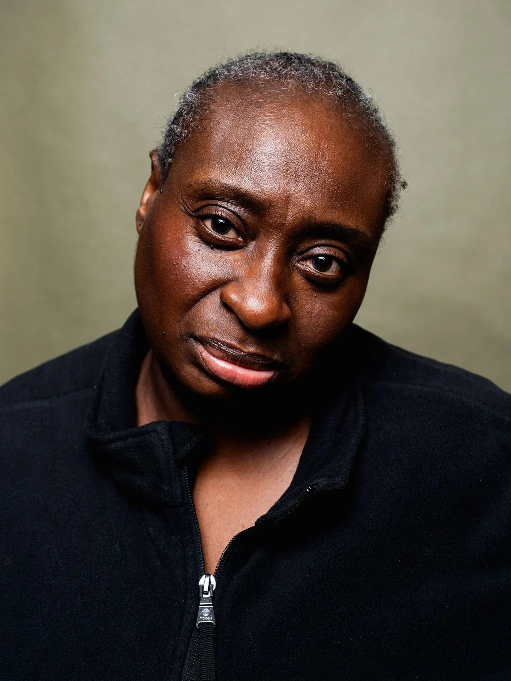 Close-up portrait of a middle-aged woman with short, grey hair, wearing a black zip-up top, looking directly at the camera against a plain background.