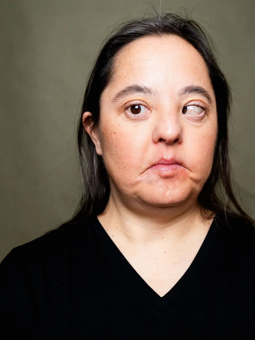 A woman with long dark hair, wearing a black shirt, looking to her left with a neutral expression.