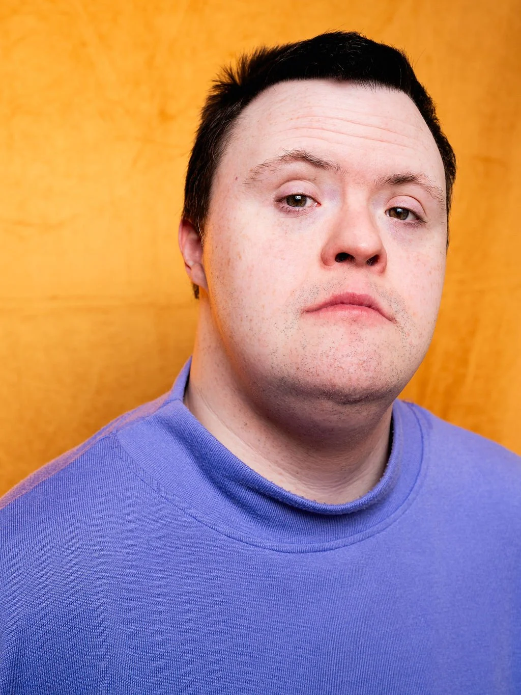 A young man with short dark hair and freckles, looking slightly at the camera, wearing a purple shirt against a yellow background.