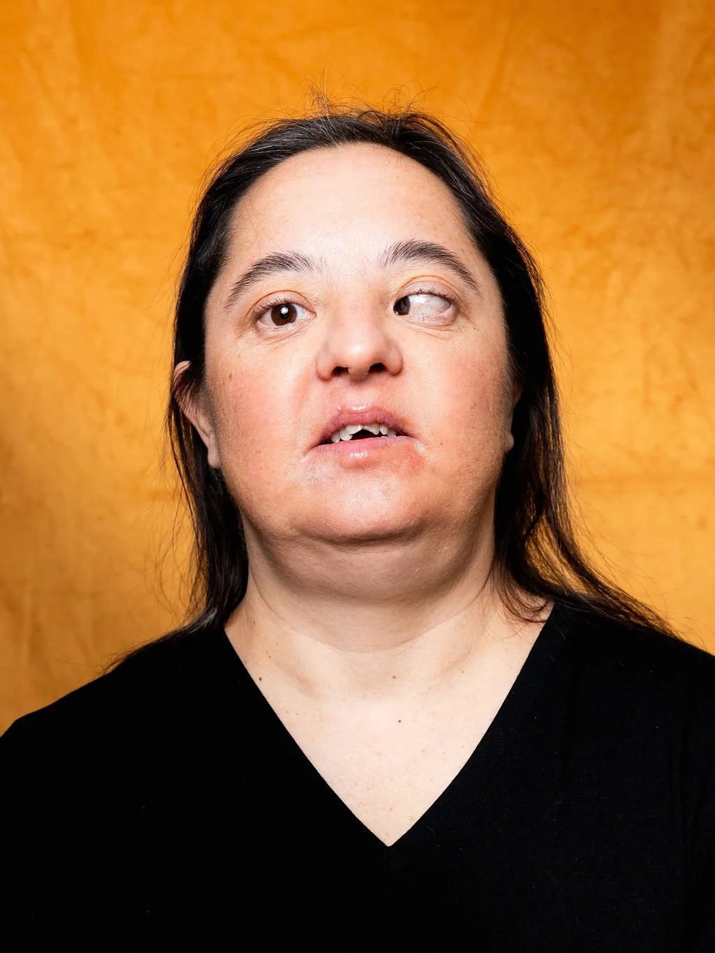 A woman with dark hair wearing a black V-neck shirt, standing against a wooden background, with her head slightly tilted and eyes looking in different directions.