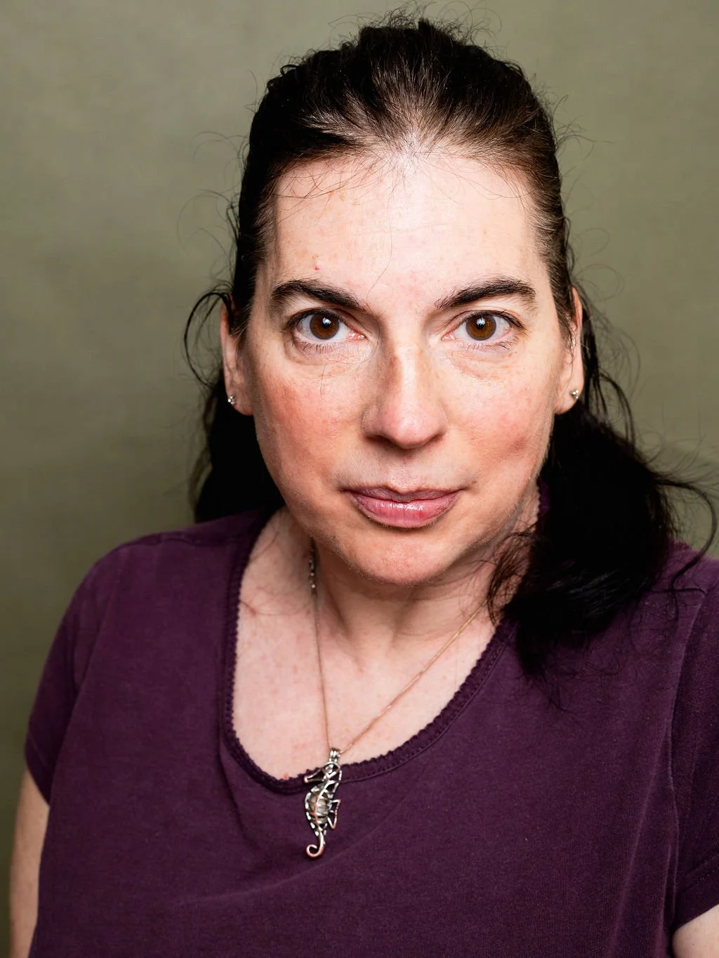 Close-up portrait of a White woman with dark hair, brown eyes, and wearing a purple top and a seahorse necklace, looking directly at the camera against a plain background.