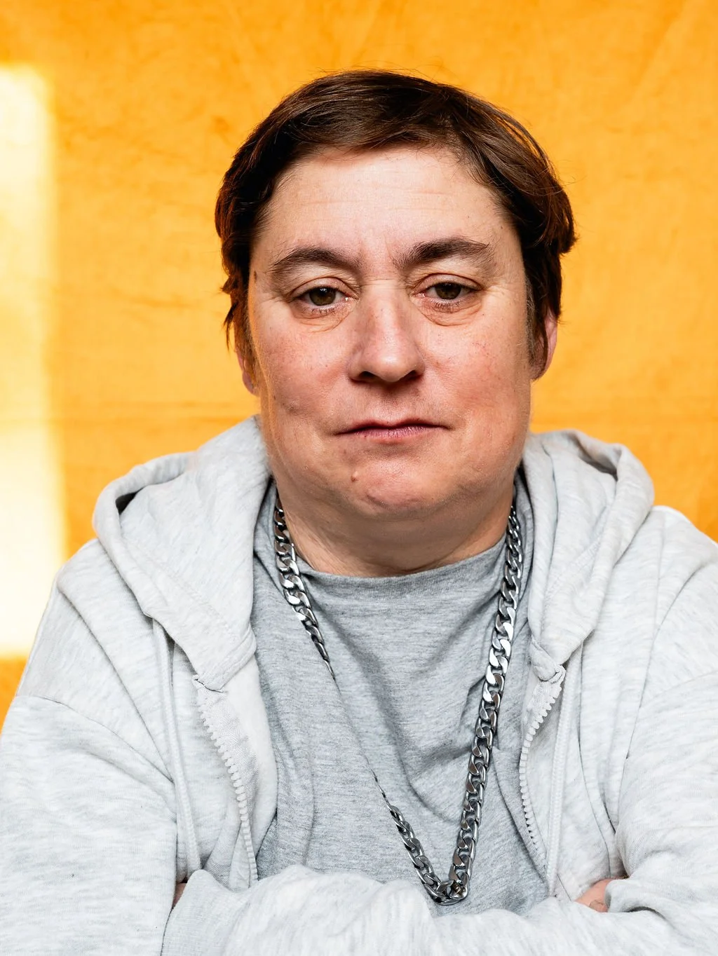 A man with short dark hair, wearing a gray hoodie, gray T-shirt, and a thick silver chain necklace, sitting against a yellow background.