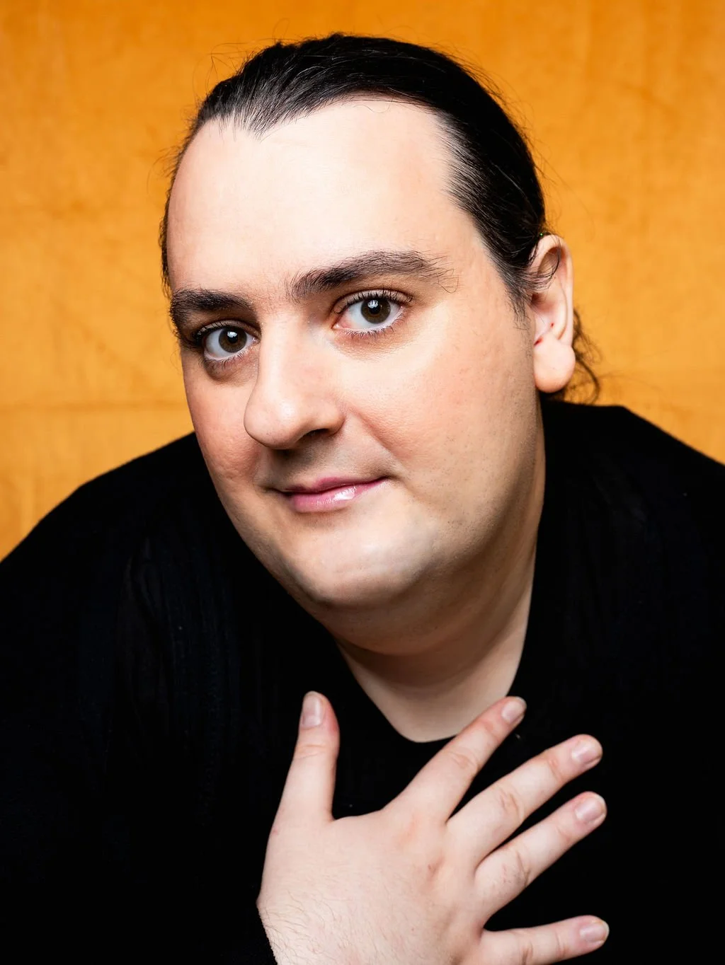 Close-up portrait of a trans woman with long dark hair, brown eyes, and wearing a black shirt, standing against a wooden background.