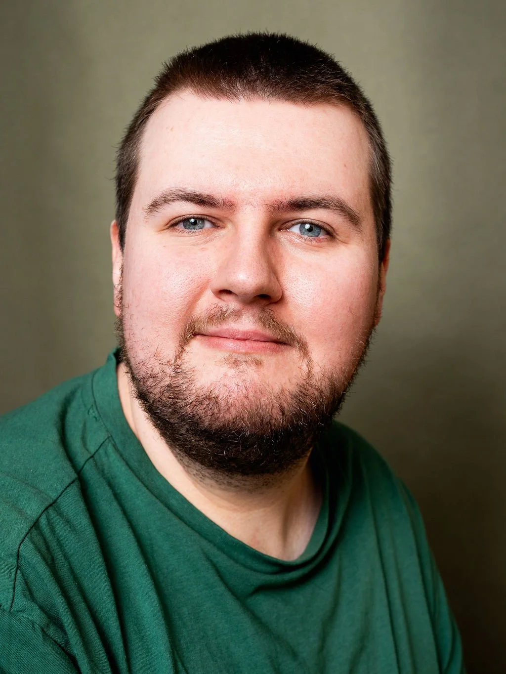 A portrait of a man with short dark hair, a beard, light blue eyes, wearing a green shirt, against a neutral background.