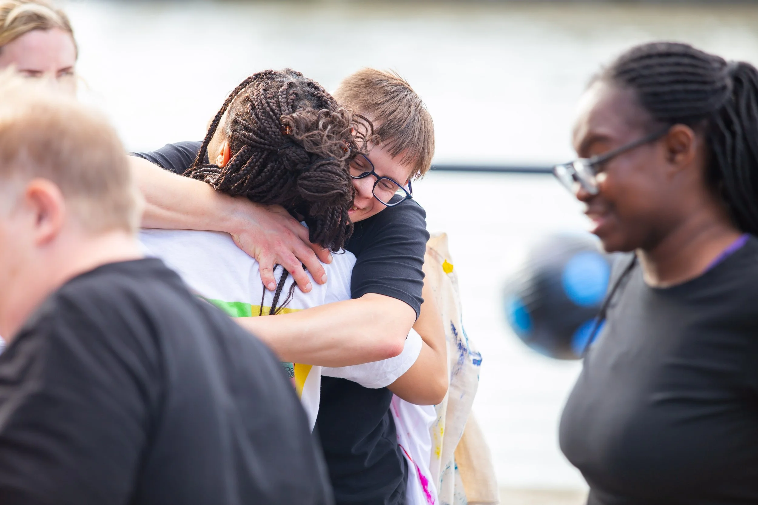 People hugging outdoors, one person wearing glasses, others smiling, some with braid hairstyles, casual clothing, in a joyful scene.