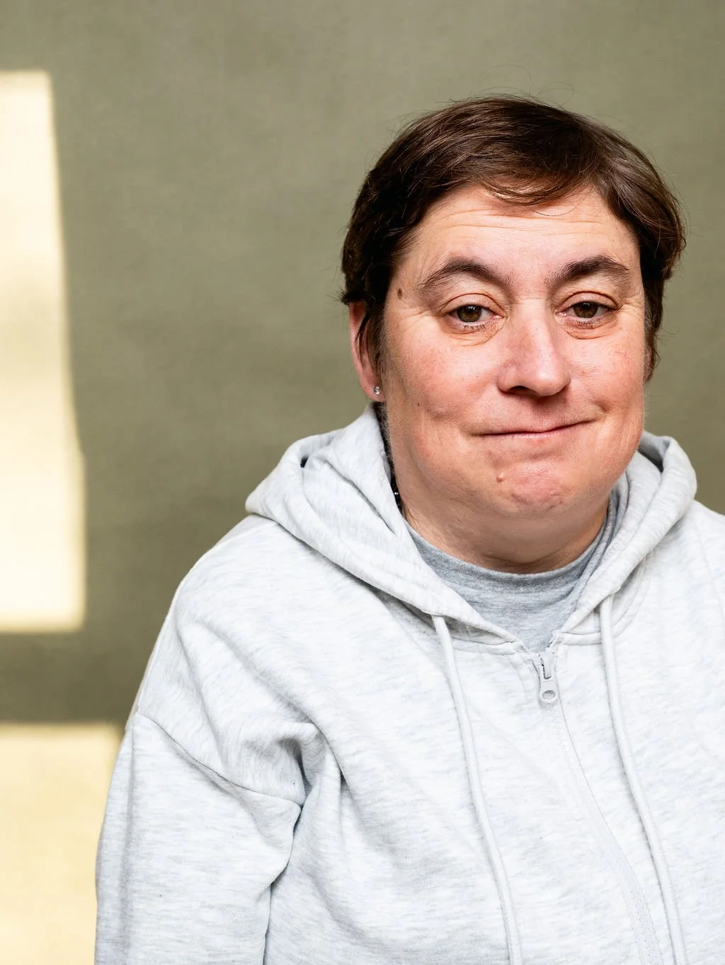 A person with short brown hair, wearing a light gray hoodie, looking slightly to the side with a neutral expression, in an indoor setting with a plain background.