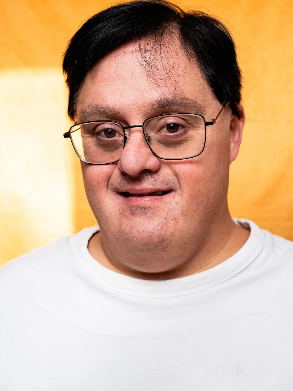 Close-up portrait of a man with black hair, glasses, wearing a white T-shirt, against a yellow background.