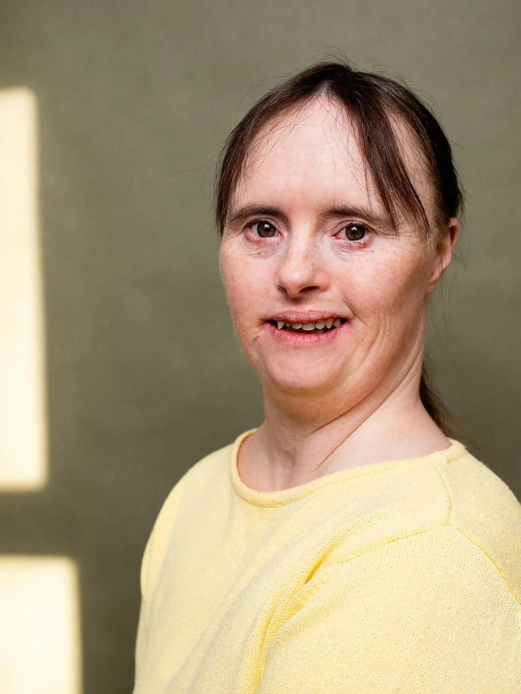 A woman with brown hair and freckles wearing a yellow shirt standing against a green background.