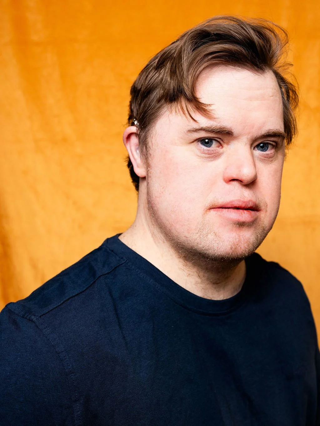 A young man with blue eyes, and brown hair styled to one side, wearing a black T-shirt, standing against a yellow background.