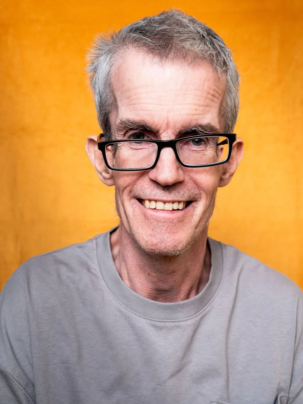 A smiling middle-aged man with grey hair, wearing black glasses and a light grey t-shirt, in front of an orange background.