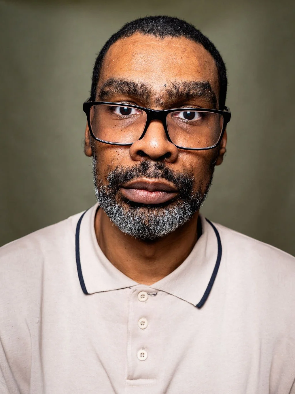 Close-up portrait of a middle-aged African American man with onyx black glasses and a gray beard, wearing a beige polo shirt with black trim, standing against a plain, muted background.