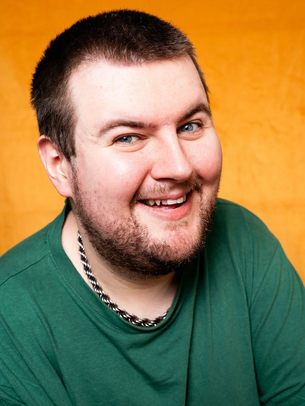 A smiling young man with short dark hair, a beard, wearing a green shirt and a striped chain necklace, against a yellow background.