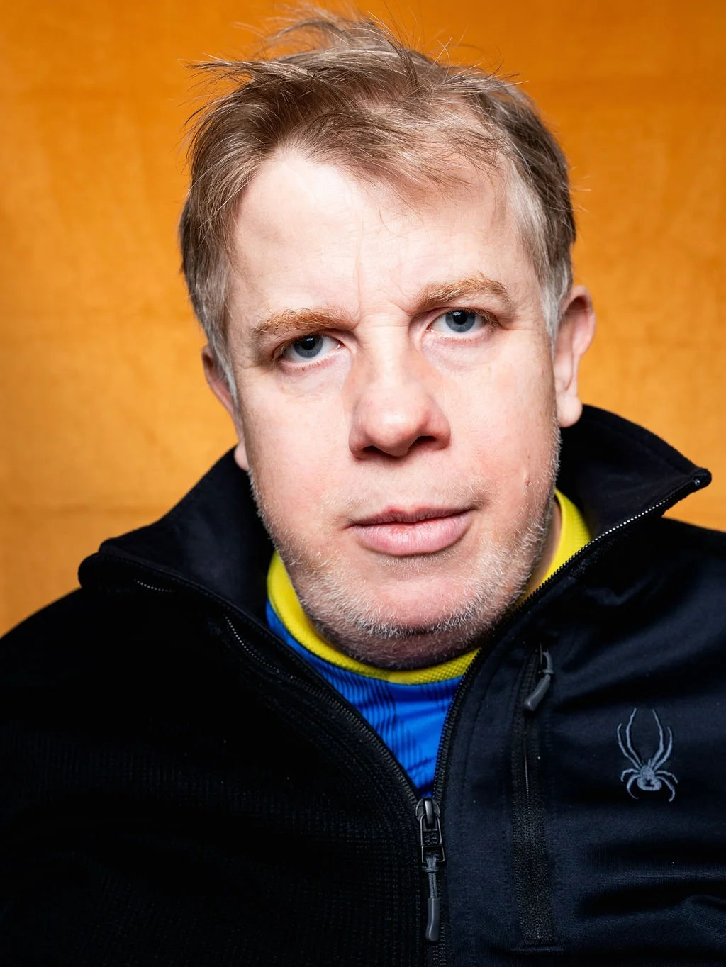 Close-up of a middle-aged man with blue eyes and light brown hair, wearing a black jacket with a spider logo, standing against a yellow background.