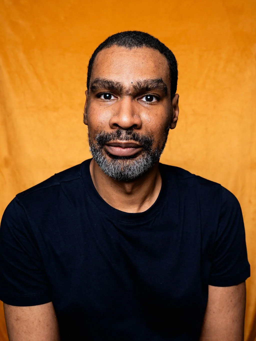 Portrait of a man with short black hair, a beard with gray, wearing a black t-shirt, against a wooden background.