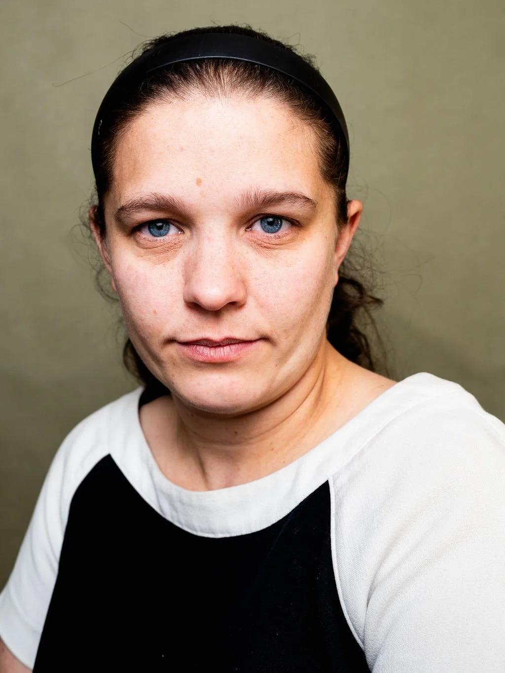 Close-up of a woman with blue eyes, dark hair pulled back with a black headband, wearing a white and black shirt against a plain background.