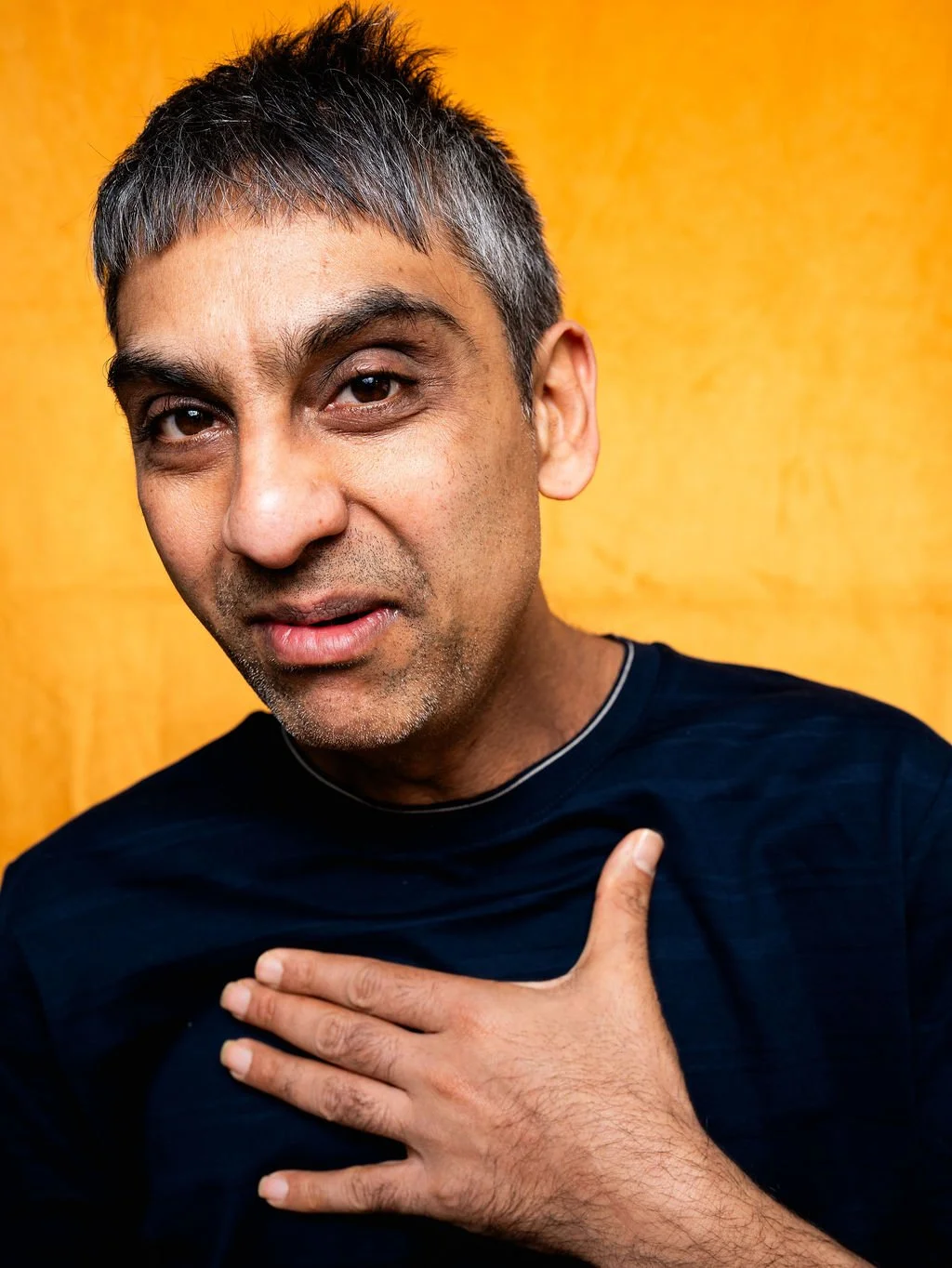 A middle-aged man with short grey hair and brown eyes making a surprised expression, with his mouth slightly open and his right hand placed on his chest, wearing a black shirt, against a yellow background.