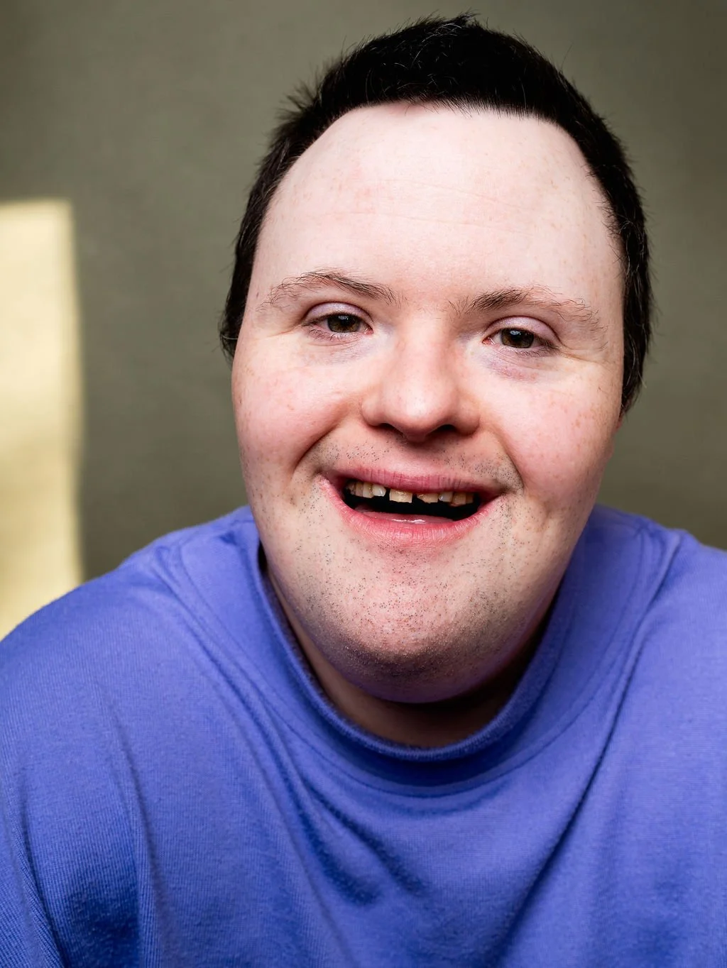 Close-up portrait of a man with short dark hair, light skin, and wearing a blue shirt, smiling, against a neutral background.