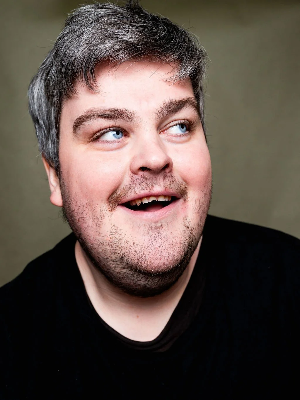 A man with short grey hair and a beard smiling and looking to his right, wearing a black shirt against a plain background.