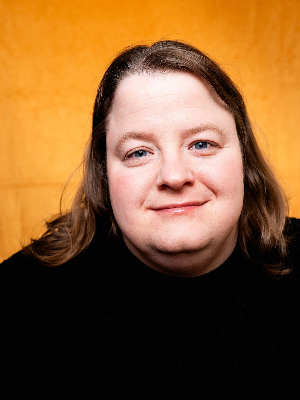 Close-up portrait of a woman with long brown hair and blue eyes, smiling, in front of a yellow background.