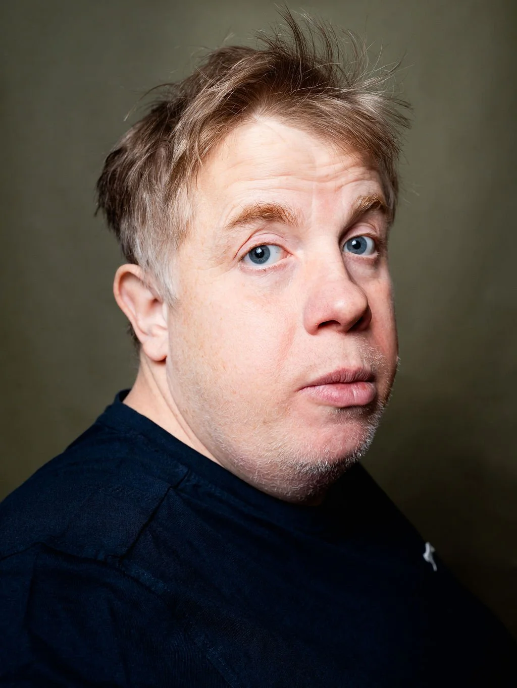 Close-up portrait of a White man with blue eyes, messy light brown hair, wearing a dark shirt, against a green background.