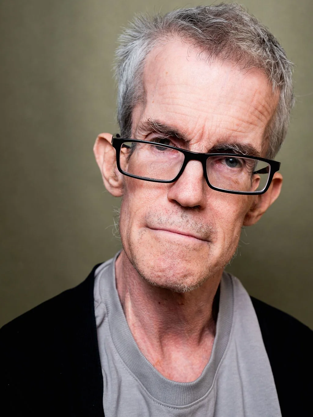 Close-up portrait of a middle-aged man with grey hair and glasses, wearing a grey shirt and black jacket against a plain background.