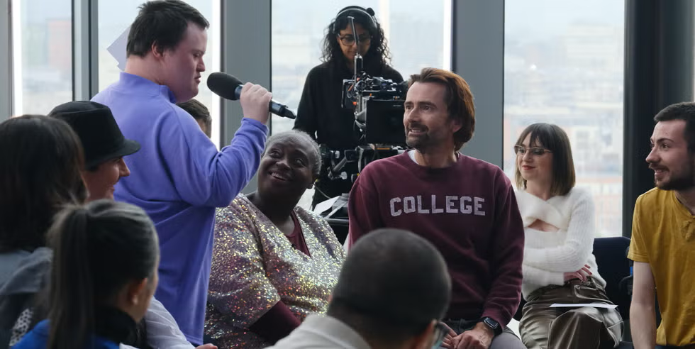 A diverse group of people participating in a discussion or interview in a modern office setting with large windows, a person with a microphone asking questions, and a camera operator recording the event.