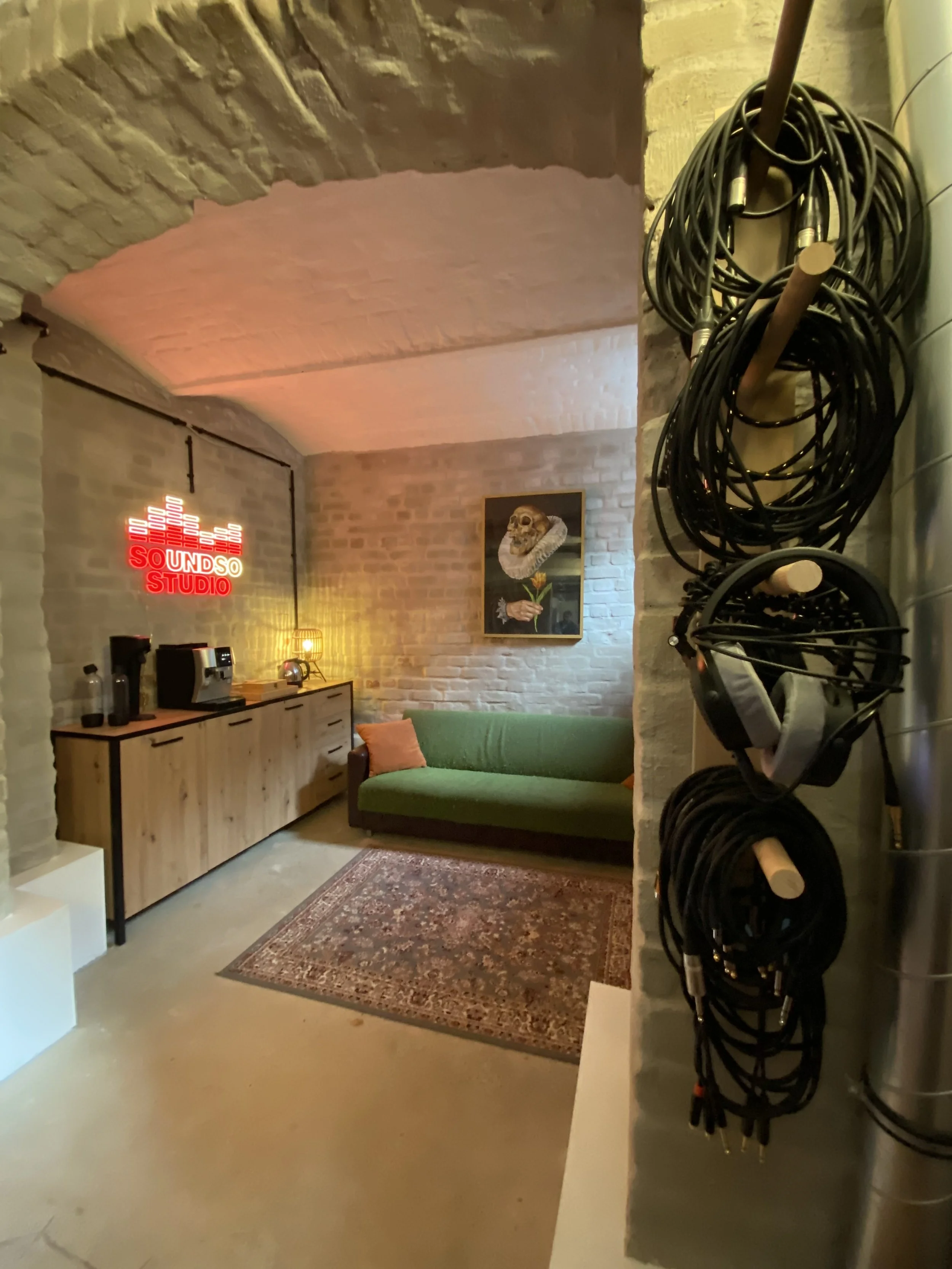 Ein gemütliches Studio mit einer grünen Couch, einem Teppich, einer Wand mit einem Kunstbild eines Schädels auf einer Rose, einer Holz-Sideboard mit Kaffeemaschine und einem Neon-Schild mit der Aufschrift 'SOUND STUDIO'.