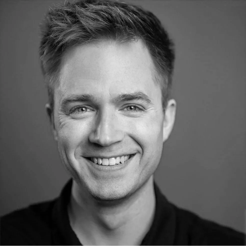 Schwarz-weiß Porträt eines lächelnden jungen Mannes mit kurzen, zerzausten Haaren, vor hellem Hintergrund.