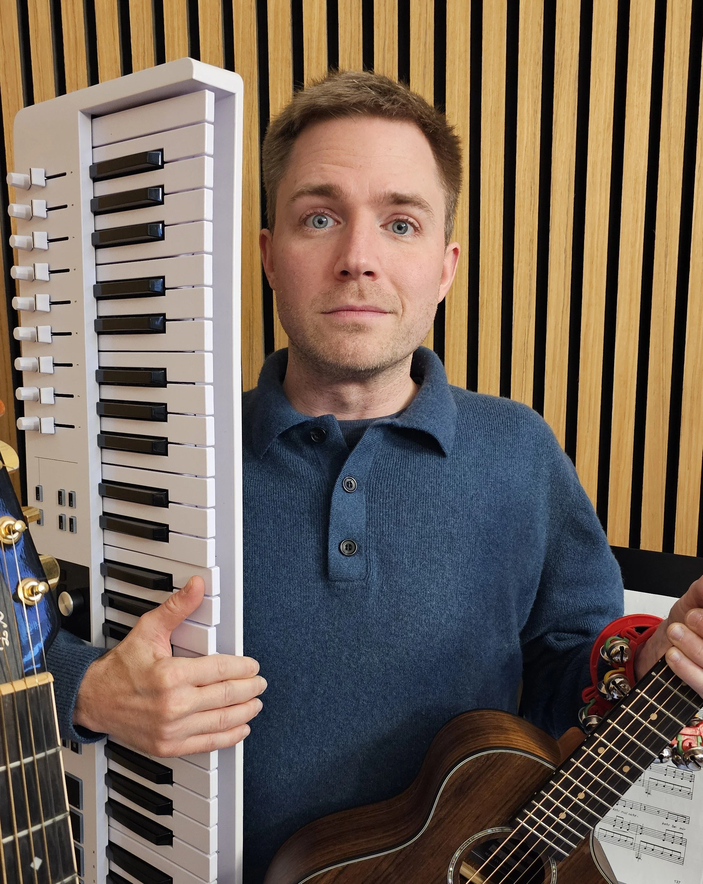 Junger Mann hält ein Keyboard und ein akustische Gitarre, steht vor einer Holzvertäfelung.