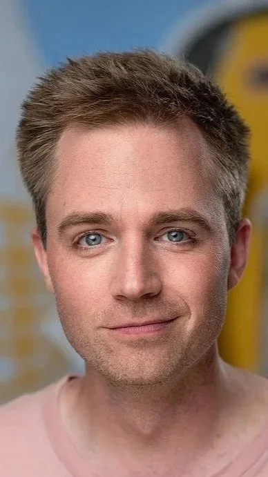 Nahaufnahme eines Mannes mit blauen Augen und braunem kurzgeschnittenem Haar vor einem gelb-blauen Hintergrund.