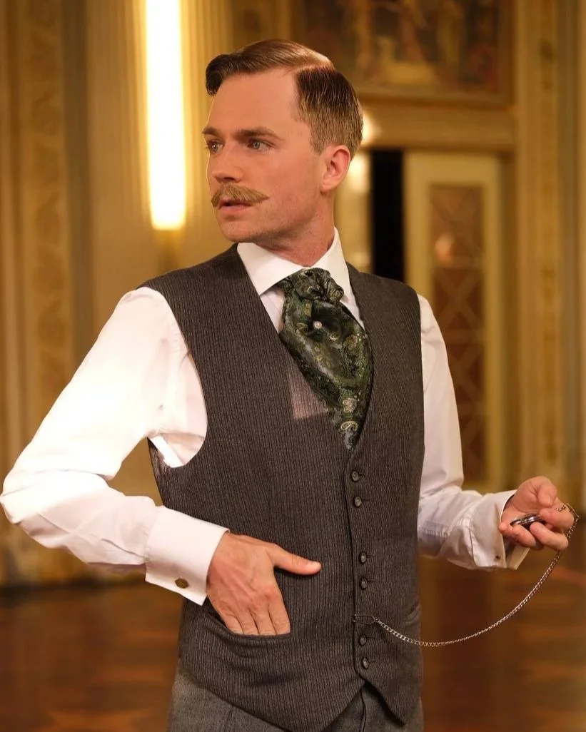 Ein Mann im viktorianischen Stil mit einem Schnurrbart, Hemd, Weste und Seidenschal, der in einem eleganten Raum steht.