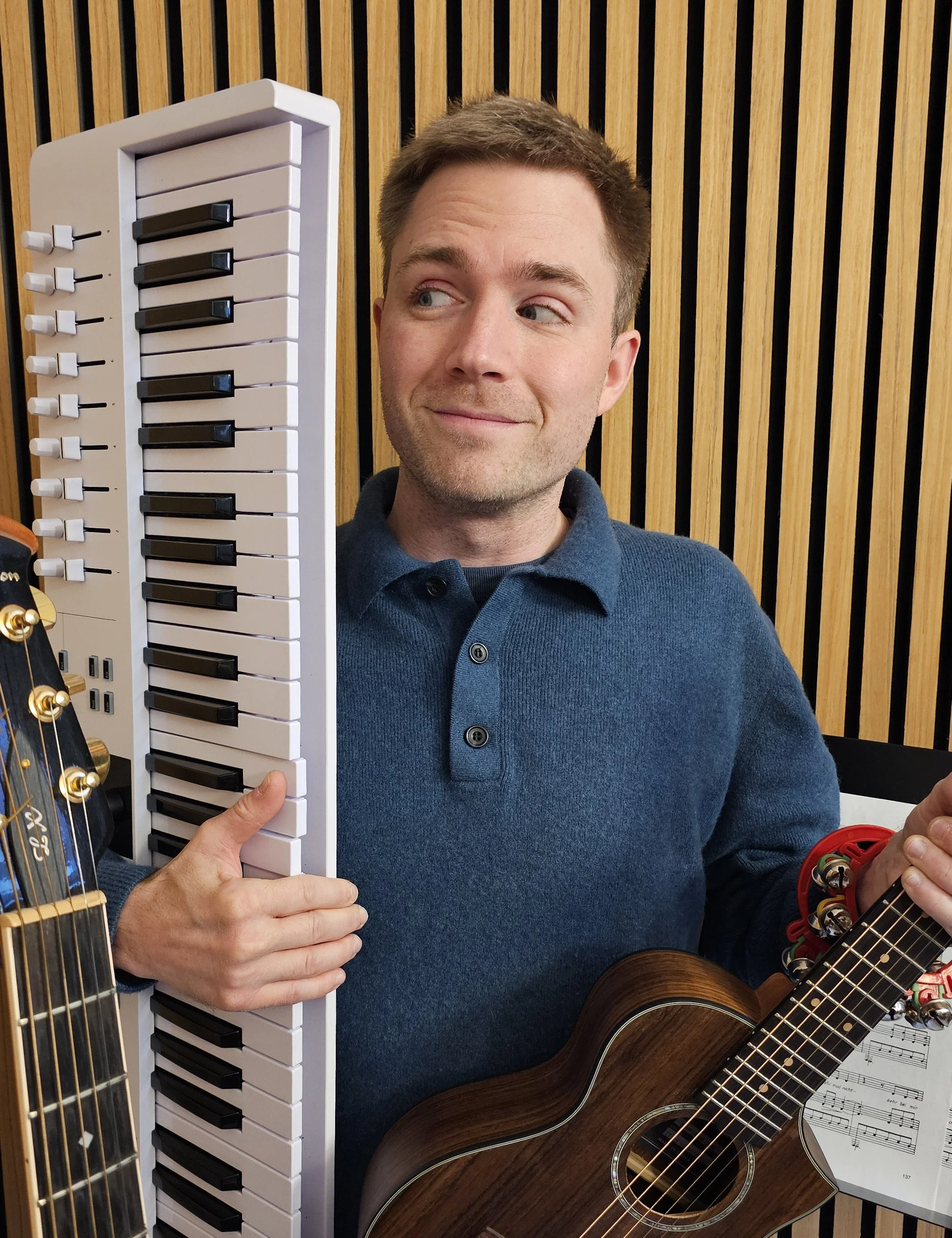 Ein Mann mit braunen Haaren, blauer Pullover, hält ein Keyboard in der Hand und steht vor einer Holzlatwand im Tonstudio