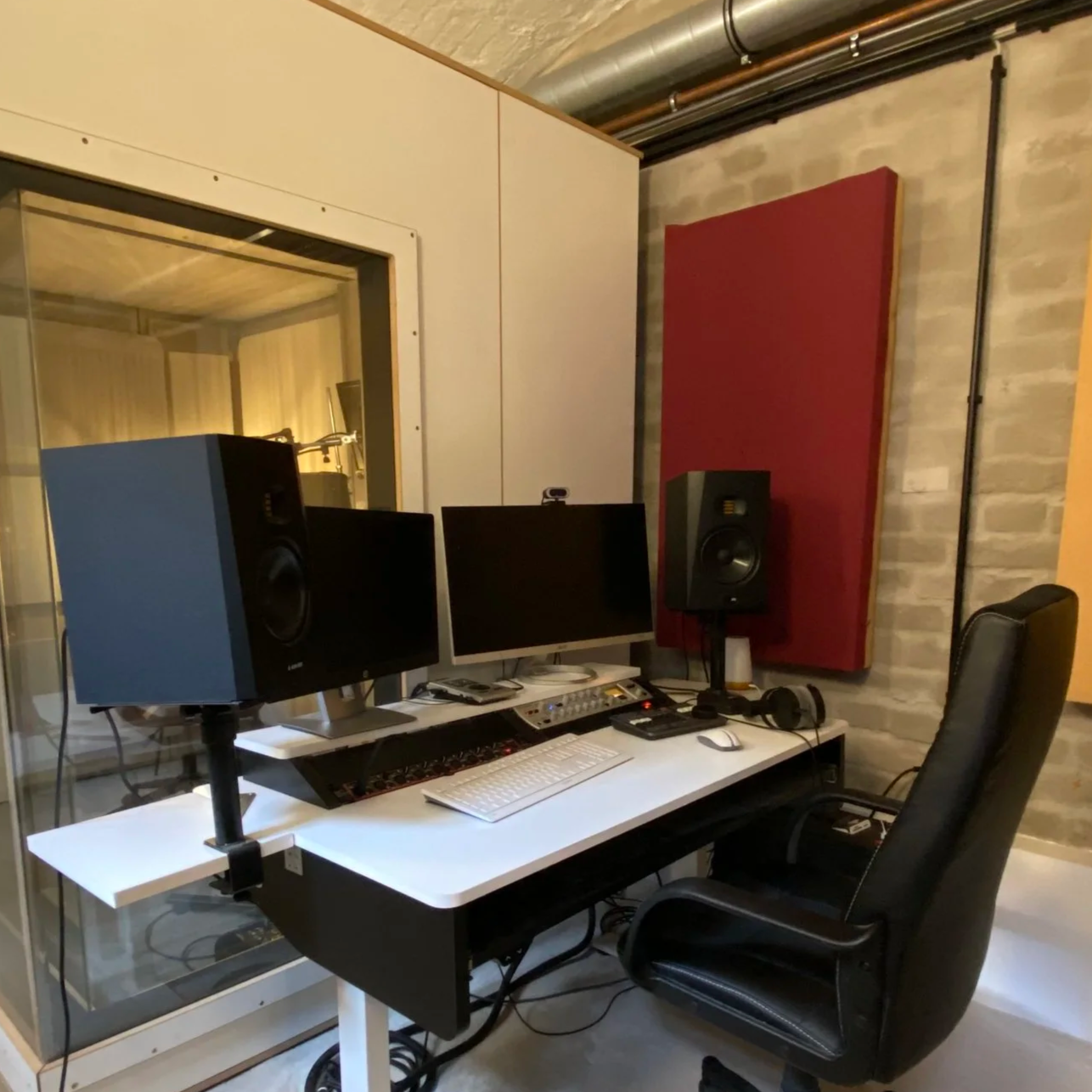Ein modernes Tonstudio mit zwei Lautsprechern, zwei Monitoren, einem Computer, einem Audio-Interface, einem Keyboard, einem Mouse, Kopfhörern, einer Klangabsorberplatte an der Wand und einem Stuhl.