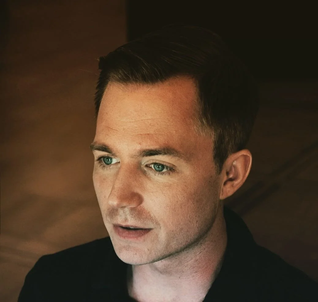 Nahaufnahme eines Mannes mit dunklen Haaren und blauen Augen, der nachdenklich schaut, vor einem dunklen Hintergrund.