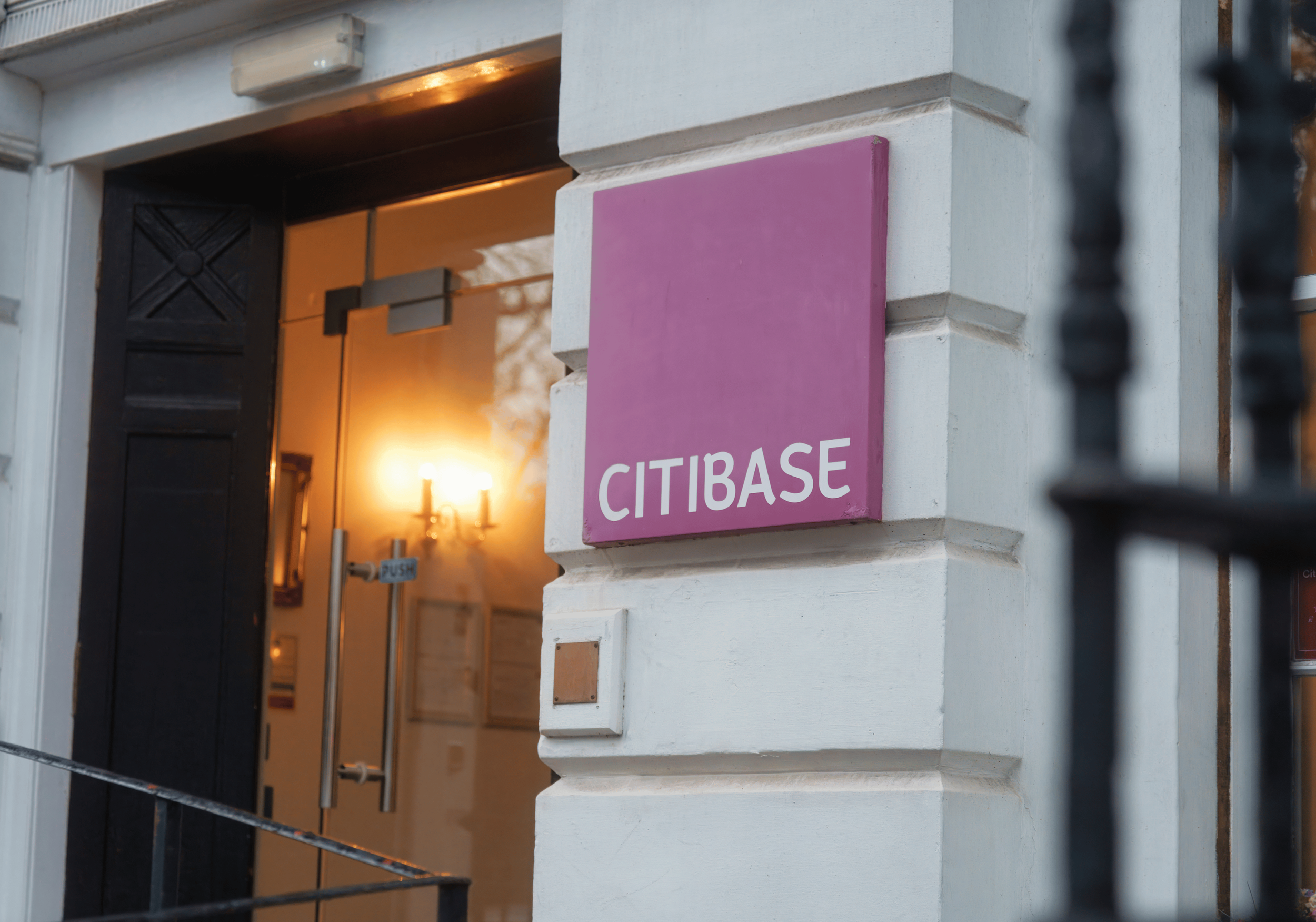 The entrance of a Citibase office building with a pink sign displaying 'CITIBASE' attached to a white brick wall, an open black door, and warm indoor lighting visible through the door.