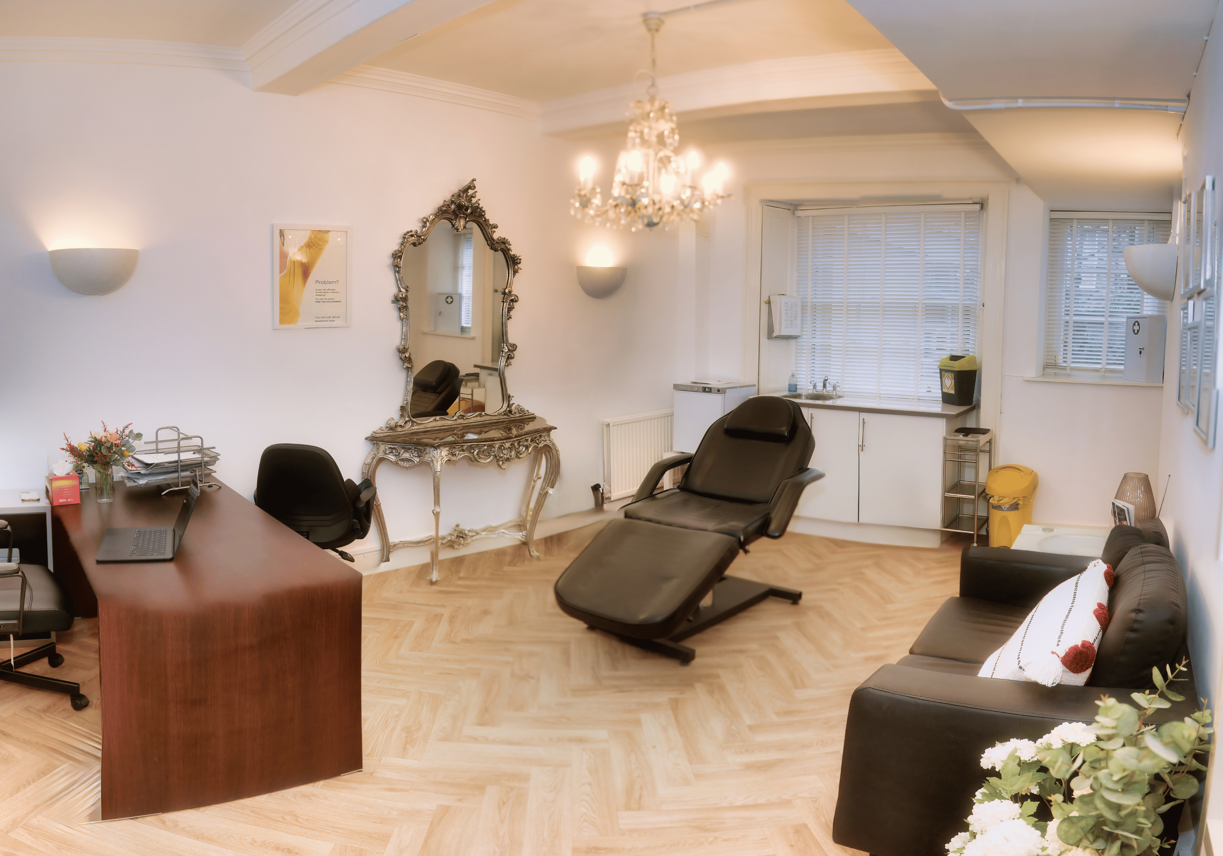 Medical consultation room with a reclining chair, black sofa with decorative pillow, wooden desk with a laptop, mirror, chandelier, and window blinds.